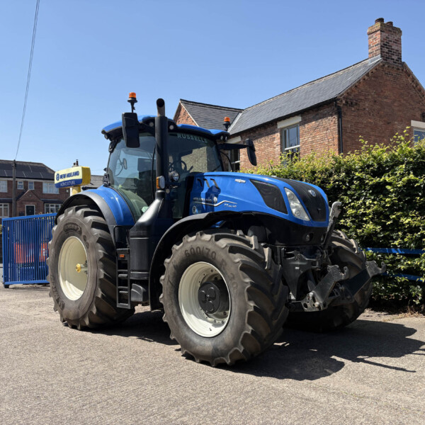 Blue New Holland tractor parked on a street in front of a brick house with a hedge and blue gate.