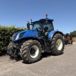 Front three-quarter view of a large blue tractor parked on a paved lot beside a green hedge and brick building against a clear blue sky at a farm equipment yard. (Informative)