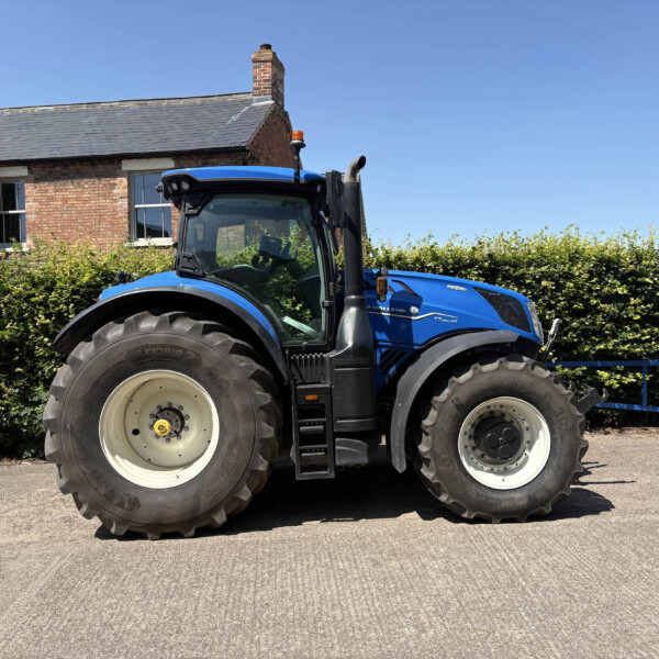Blue New Holland tractor parked on a paved driveway with a brick house and green hedge behind it under a clear blue sky, front-left wheel in view.