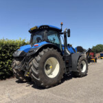 Blue agricultural tractor with large rear tires parked on a paved lot; hedge on the left and a brick building in the background under a clear blue sky.