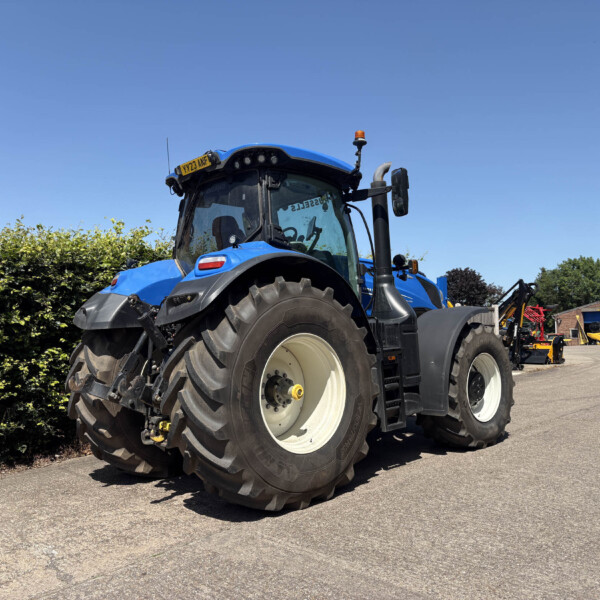 Blue agricultural tractor with large rear tires parked on a paved lot; hedge on the left and a brick building in the background under a clear blue sky.