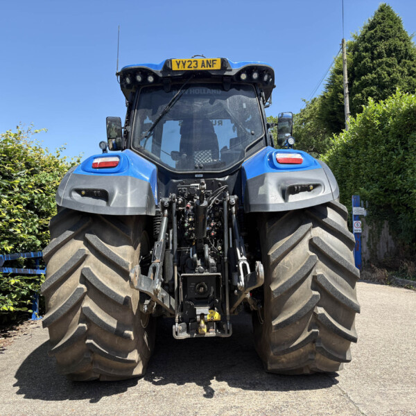 Rear view of a blue agricultural tractor with oversized tires, parked on a paved road, flanked by green hedges under a clear blue sky