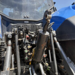 Close-up of a blue tractor’s front hydraulic system with levers, hoses, and metal fittings visible amidst dust and grime.