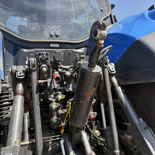 Close-up of a blue tractor’s front hydraulic system with levers, hoses, and metal fittings visible amidst dust and grime.