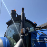 Close-up view of a blue New Holland tractor cab with a tall exhaust pipe and orange warning beacon against a clear blue sky