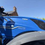 Close-up of a blue New Holland tractor cab with orange beacon and brick chimney against a blue sky forgrounded in the image context?