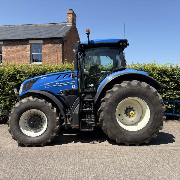 Blue New Holland tractor in front of a brick building with hedges under a clear blue sky