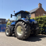 Blue tractor parked on a street beside brick houses and a blue fence under a clear blue sky? Actually the fence is blue gate—Blue tractor on a sunny urban street next to brick houses and greenery.