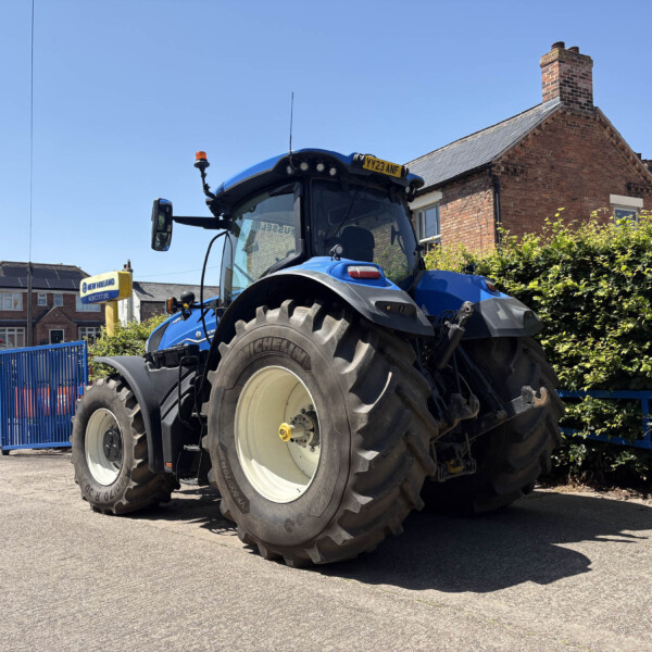 Blue tractor parked on a street beside brick houses and a blue fence under a clear blue sky? Actually the fence is blue gate—Blue tractor on a sunny urban street next to brick houses and greenery.