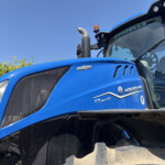 Blue New Holland tractor close-up of cab and exhaust against clear sky, with brick building and greenery in background