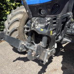 Close-up of a blue tractor's front end, showing a large tire, hitch, and hydraulic components with warning stickers.
