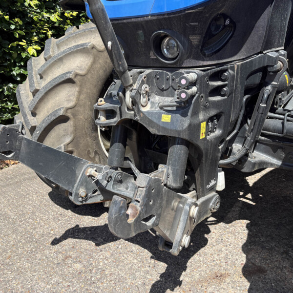 Close-up of a blue tractor's front end, showing a large tire, hitch, and hydraulic components with warning stickers.