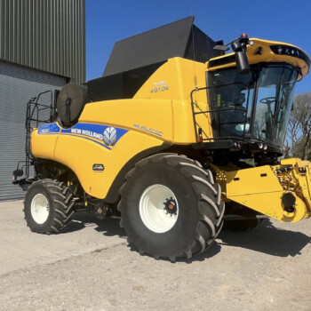 Front-right view of a bright yellow New Holland combine harvester with oversized tires, parked beside a gray metal building against a clear blue sky.