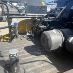 Industrial machinery mounted on a flatbed truck with hoses, ducts, and exposed wiring visible near large tires.