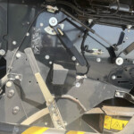 Close-up of a dark metal machine frame with bolts, hoses, and warning labels visible on the surface.