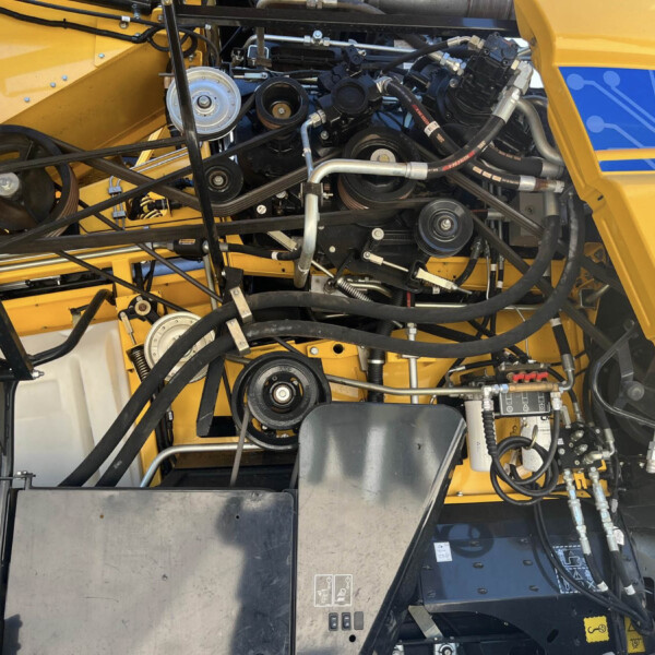 Complex engine bay of a yellow industrial machine with belts, hoses and pulleys exposed.