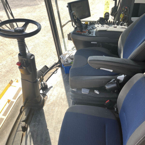 Interior of a tractor cab with a steering wheel on the left and a control console with buttons and a screen in the center.