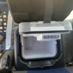 Inside a vehicle cab showing a black seat belt, a grey vent, and a metal footwell with a perforated step nearby; industrial equipment visible on left side.