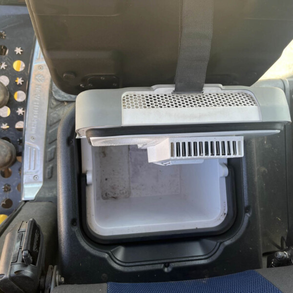 Inside a vehicle cab showing a black seat belt, a grey vent, and a metal footwell with a perforated step nearby; industrial equipment visible on left side.