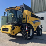 Yellow New Holland combine harvester parked beside a metal-clad building under a clear blue sky, showing front header and large black tires.