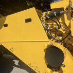 Close-up of yellow construction machine frame showing hydraulic hoses, bolts, and warning decals on metal panels.