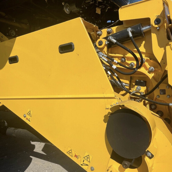Close-up of yellow construction machine frame showing hydraulic hoses, bolts, and warning decals on metal panels.