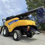 New Holland yellow combine harvester with a long gray unloading tube and a blue tarp-covered end, parked in a lot under a cloudy sky.