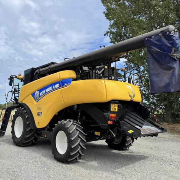 New Holland yellow combine harvester with a long gray unloading tube and a blue tarp-covered end, parked in a lot under a cloudy sky.