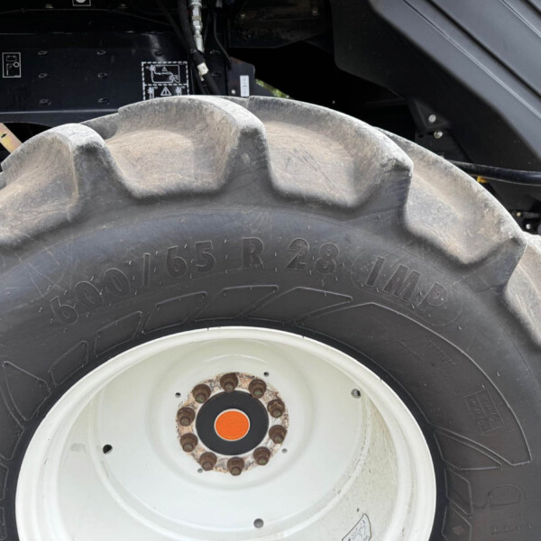 Close-up of a large dusty tractor tire with a white rim and rusted lug bolts, part of farming equipment behind it.