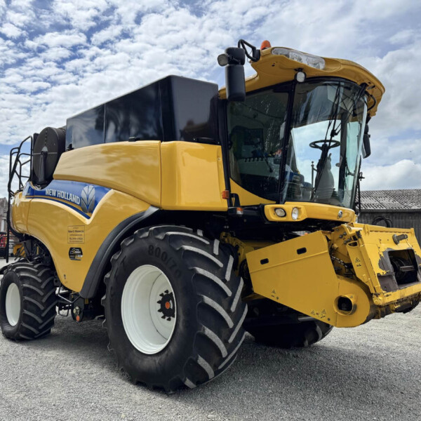 Front-right view of a large yellow New Holland combine harvester with oversized white wheels parked in a lot, blue logo visible on the body.