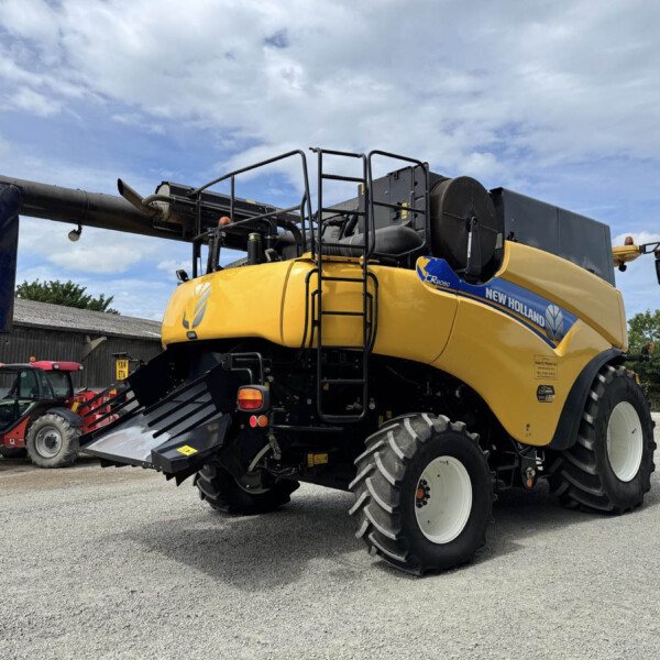 New Holland CR8080 combine harvester in yellow with a large black header, parked outdoors on gravel under a cloudy sky.
