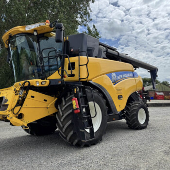 New Holland yellow combine harvester photographed from the front-left, parked on gravel with trees and buildings in the background.