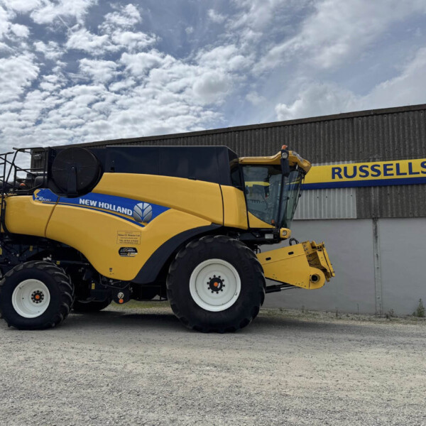 Yellow New Holland combine harvester parked on gravel in front of a gray industrial building with a blue and yellow Russell's sign on the wall