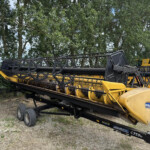 Yellow and black agricultural harvester header mounted on a trailer, with a tree-lined background; outdoors on a gravel surface.