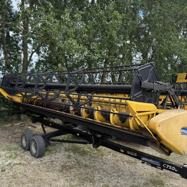 Yellow and black agricultural harvester header mounted on a trailer, with a tree-lined background; outdoors on a gravel surface.