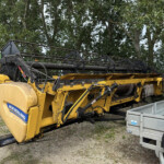 Yellow New Holland agricultural header attached to a trailer in a yard, with trees in the background