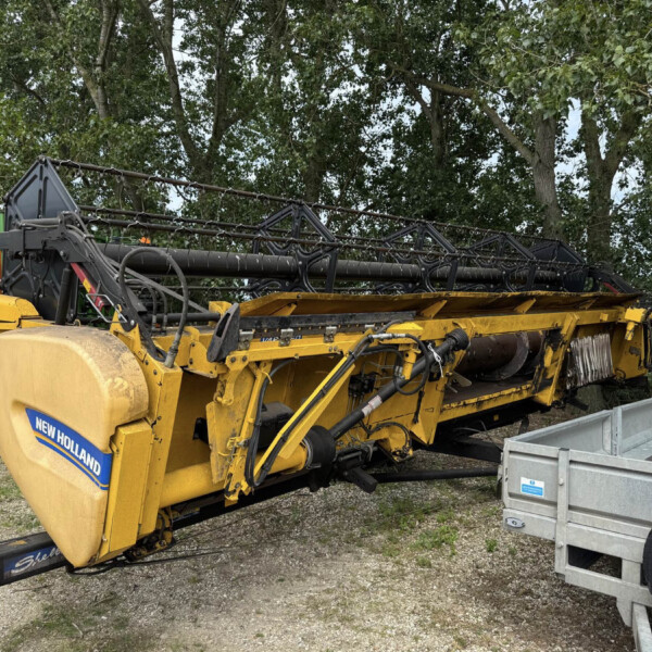 Yellow New Holland agricultural header attached to a trailer in a yard, with trees in the background
