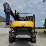 Rear view of a yellow excavator with a large blue-covered arm, parked on a gravel lot near a corrugated metal building.