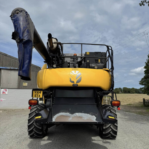 Rear view of a yellow excavator with a large blue-covered arm, parked on a gravel lot near a corrugated metal building.