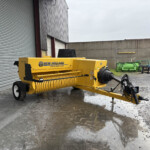 Yellow New Holland Hayliner 275 baler parked beside a corrugated metal building on a wet gravel lot with other green farm equipment in the background.