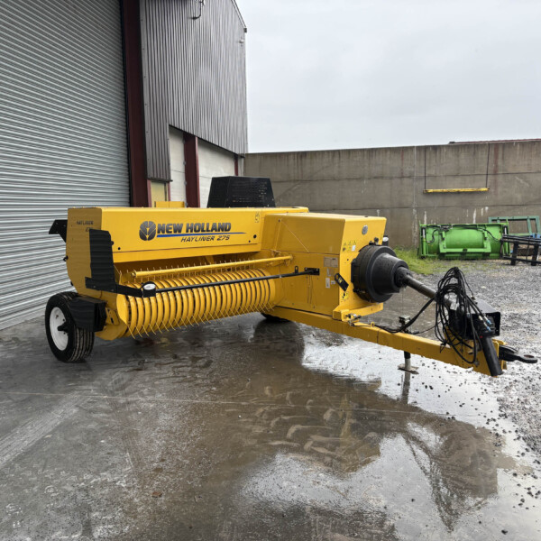 Yellow New Holland Hayliner 275 baler parked beside a corrugated metal building on a wet gravel lot with other green farm equipment in the background.