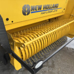 Close-up of a yellow New Holland Hayliner 275 forage header with curved pick-up tines and metal guards on a farm field machine