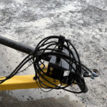 Close-up of hydraulic hoses and cables on a yellow construction machine, with a muddy, gravel-covered ground in the background.