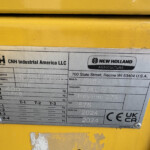Metal identification plate mounted on yellow equipment, showing CNH Industrial America LLC and New Holland Agriculture branding with model/destination fields.