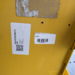Yellow metal surface with two white labels, a visible QR code, and a barcode; some peeling paint and a small hole indicate wear.