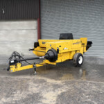 Yellow New Holland agricultural machine parked in front of a large gray corrugated metal door in an industrial setting, with wet, muddy ground nearby.