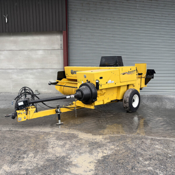 Yellow New Holland agricultural machine parked in front of a large gray corrugated metal door in an industrial setting, with wet, muddy ground nearby.