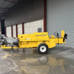 Yellow New Holland Hayliner baler parked beside a large corrugated metal warehouse with a rolling shutter, on wet muddy ground after rain still drifting moisture in the air.