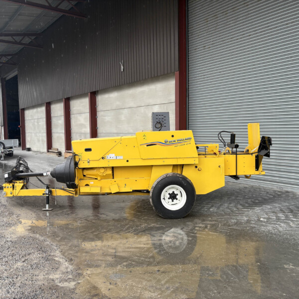 Yellow New Holland Hayliner baler parked beside a large corrugated metal warehouse with a rolling shutter, on wet muddy ground after rain still drifting moisture in the air.