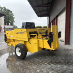 Yellow New Holland Hayliner 275 baler parked inside a metal-shed storage area on a wet concrete floor with large rolling doors nearby